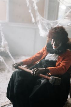 一个令人毛骨悚然的小丑穿着服装，坐在幽灵般的房间里，手里拿着一把被蜘蛛网包围的锤子