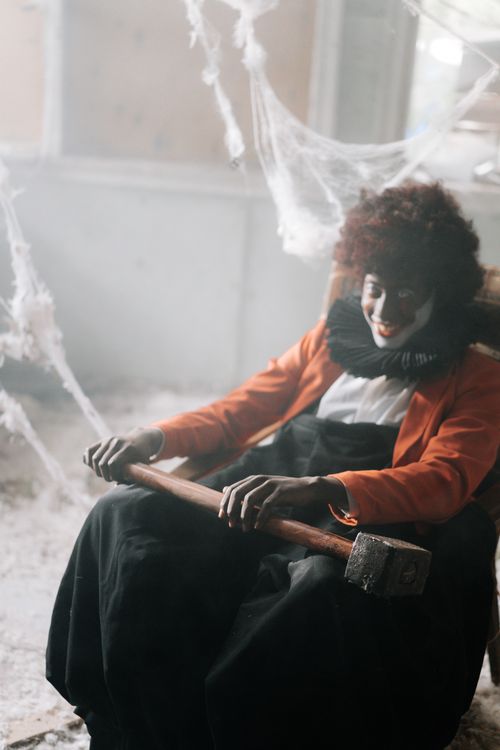 Free A creepy clown in costume sitting in a spooky room holding a hammer surrounded by cobwebs. Stock Photo
