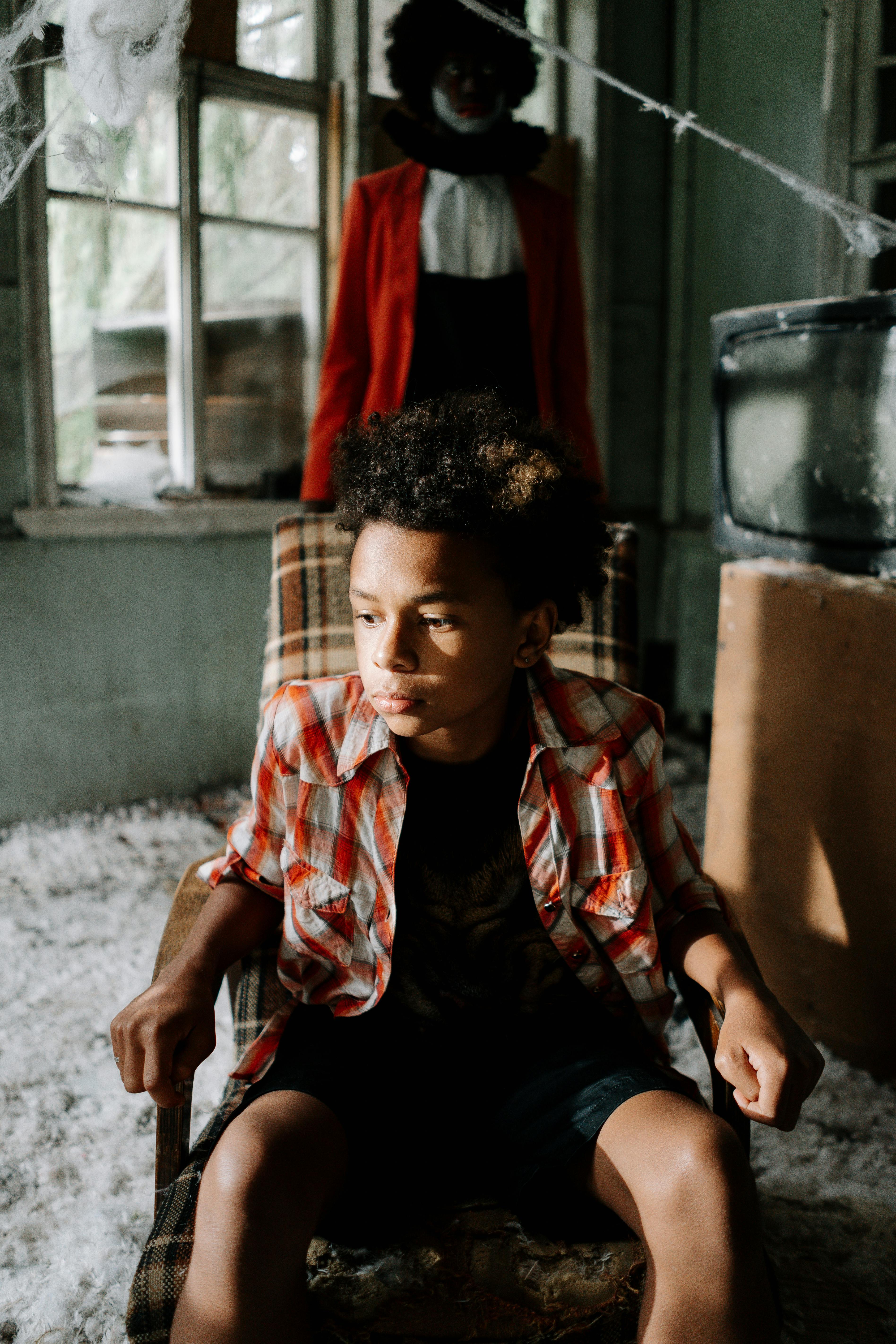 昏暗的房间里，一个穿着格子衬衫的小男孩坐在椅子上，身后不祥地站着一个幽灵般的小丑