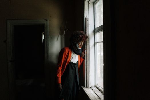 A person in a clown costume ominously gazes out a dimly lit window inside an abandoned building.