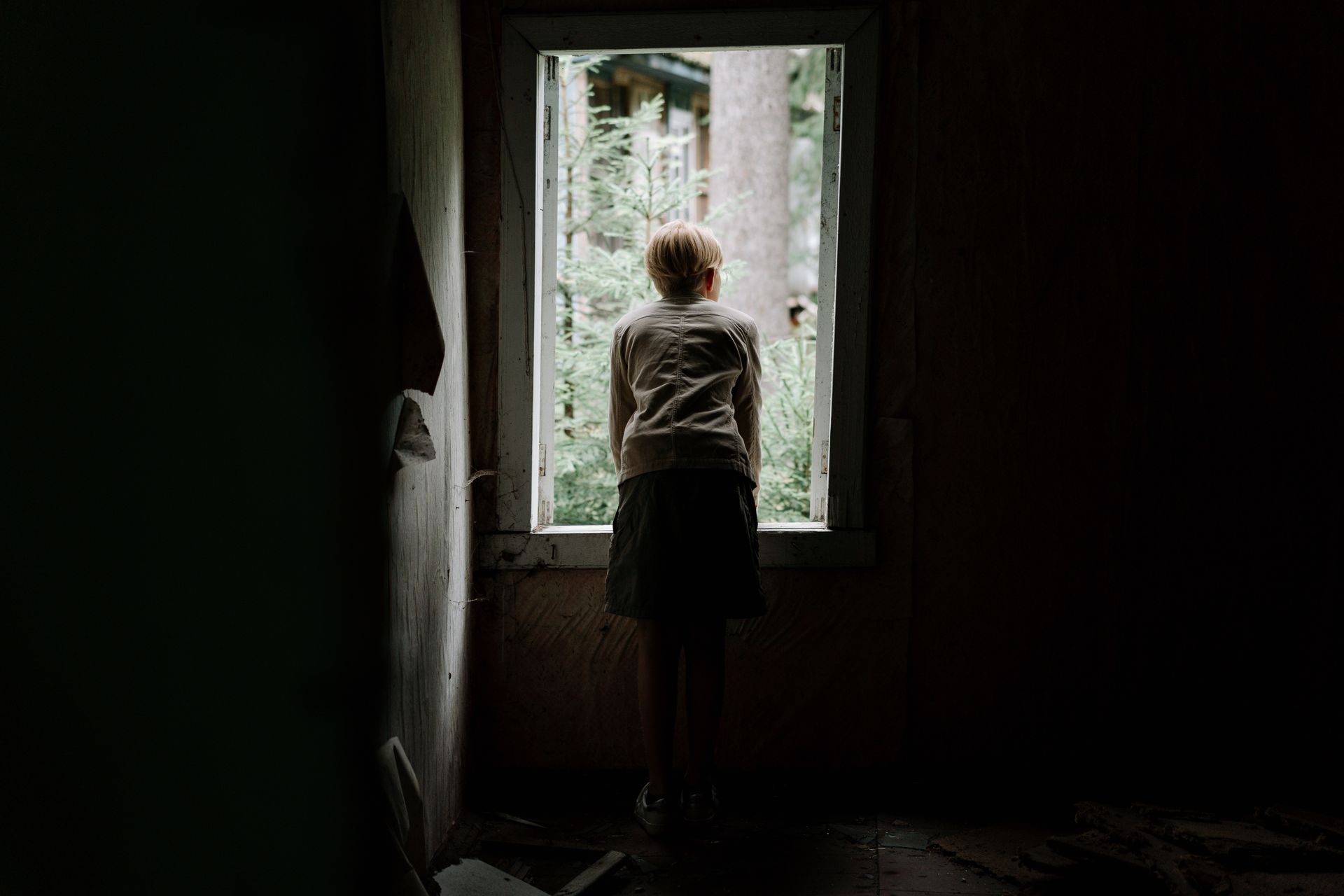 A young child curiously looks through a window of an abandoned building into the greenery outside.