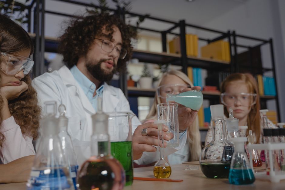 Diverse students in protective goggles study chemistry with a teacher, conducting experiments in a classroom setting.
