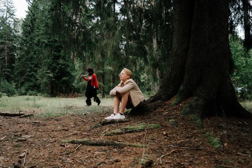 Free A teenager sits by a tree in a forest, unaware of a masked figure approaching. Stock Photo