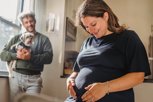 Cheerful adult pregnant woman feeling child moving while touching belly and husband smiling while standing with dog near window in apartment