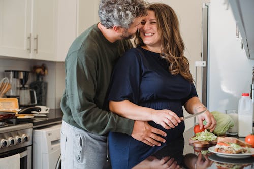 Free Cheerful pregnant wife in blue dress cooking sandwiches for breakfast while smiling and looking at husband embracing belly in modern kitchen Stock Photo