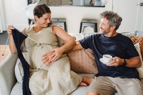 From above of smiling adult bearded man sitting on couch with hot coffee and touching pregnant belly of happy wife with knitting needles