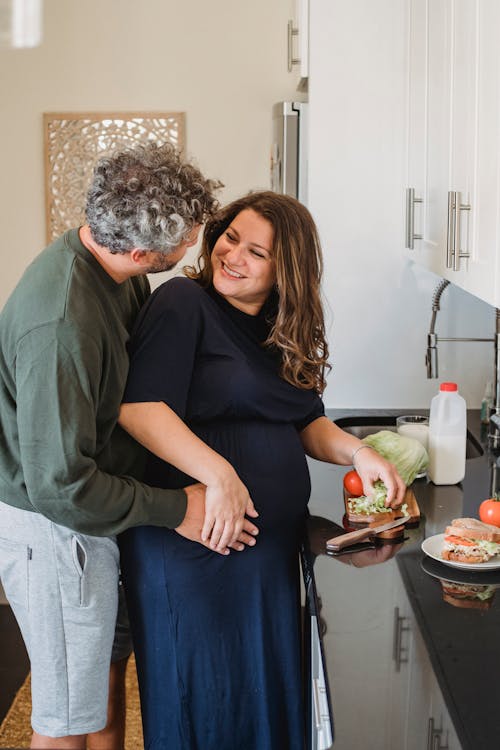 Positive pregnant woman looking at husband while cooking sandwiches