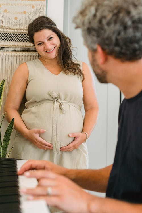 Crop anonymous adult man playing song on piano for pregnant wife in casual dress smiling and standing near wall at home