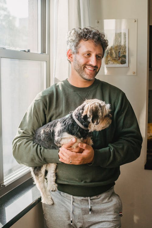 Free Happy adult man holding purebred dog while standing near window Stock Photo