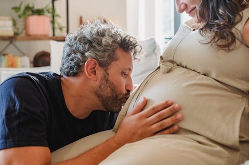 Side view of loving adult husband gently touching and kissing pregnant tummy of crop wife sitting on couch in light apartment