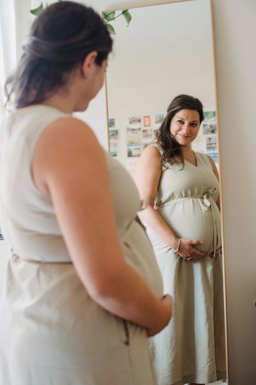 Free Happy adult lady in domestic dress touching pregnant belly while standing in light apartment and looking in mirror Stock Photo