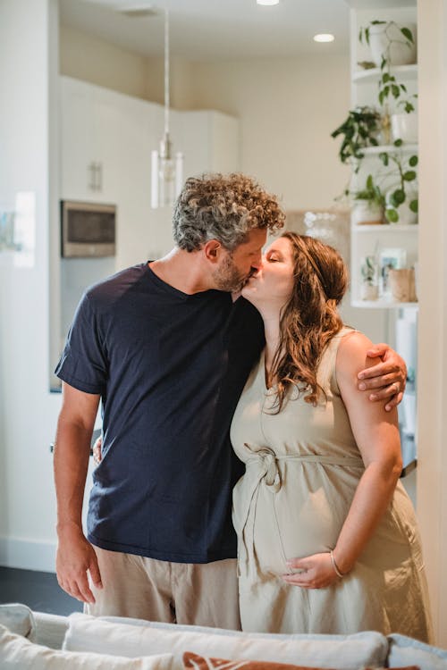 Happy adult pregnant wife and husband kissing while standing behind couch