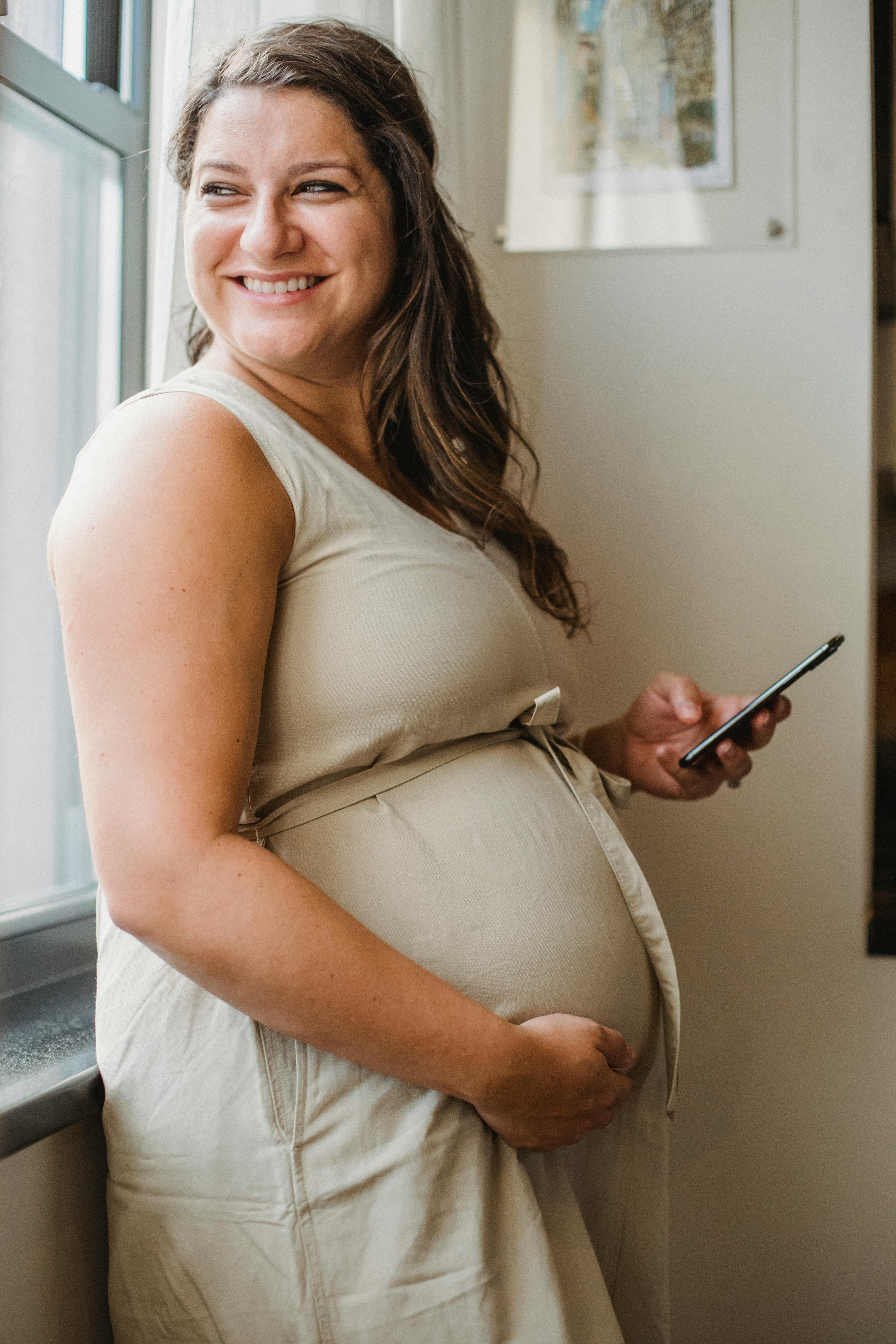 happy pregnant woman with phone at window