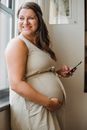 Side view of laughing woman touching belly while looking away in window and browsing smartphone at home