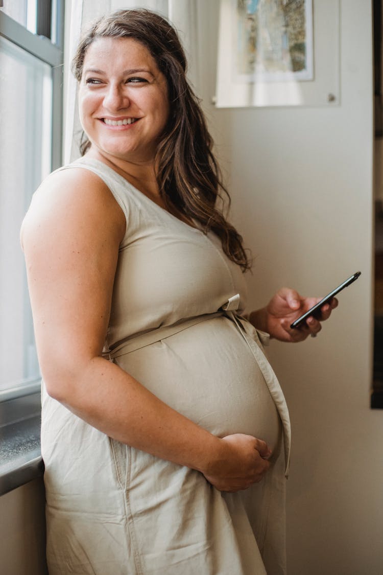 Happy Pregnant Woman With Phone At Window