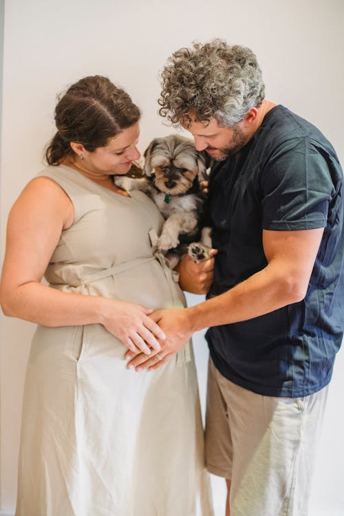 Smiling pregnant couple embracing dog and touching tummy near wall