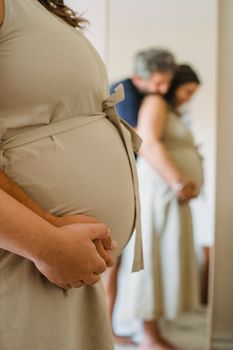 灯光室里，一名无法辨认的男子的侧面图站在镜子前，拥抱并触摸怀孕妻子的肚子