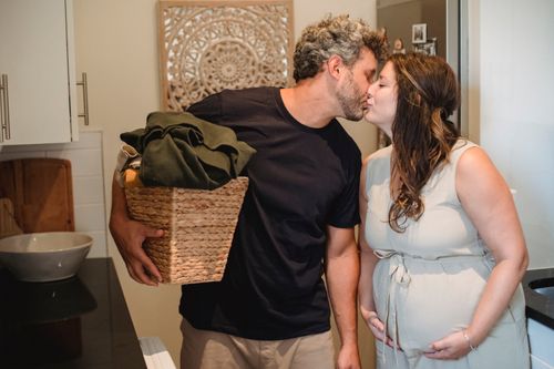 Free Happy adult man with laundry basket in hand kissing pregnant wife with closed eyes while standing together in bathroom Stock Photo