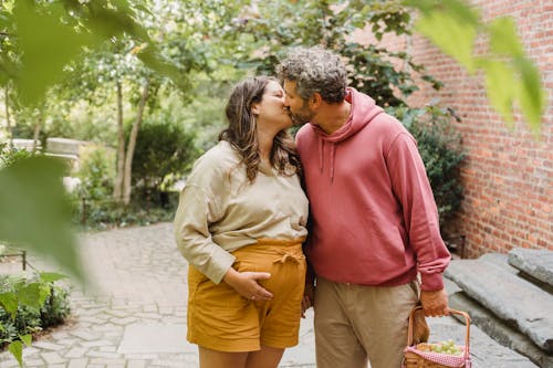 Free Happy pregnant woman in casual clothes touching tummy while kissing adult husband holding basket with fresh fruits during holidays in countryside Stock Photo