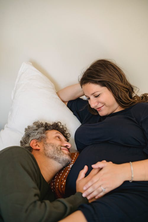 High angle of happy bearded adult man with gray hair touching tummy of regnant wife while lying together on comfortable bed and looking at each other