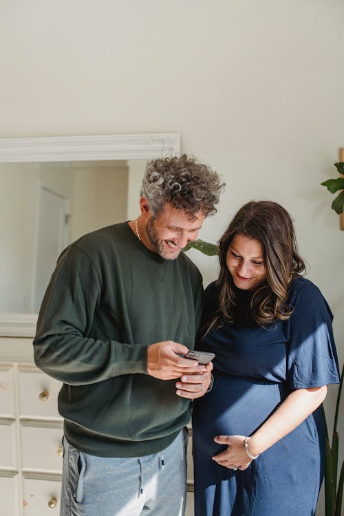 Cheerful adult male in casual clothes smiling and showing video on smartphone to smiling pregnant wife standing in light room on sunny day