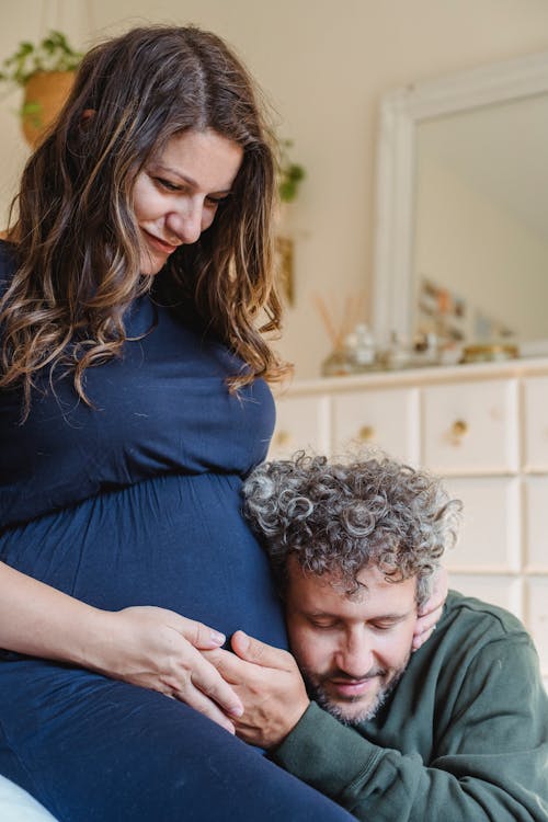 Happy adult man with closed eyes listening to belly of pregnant wife sitting on bed in cozy room