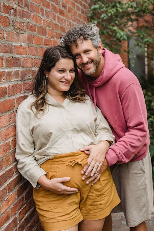 Smiling pregnant couple cuddling and touching tummy near brick house