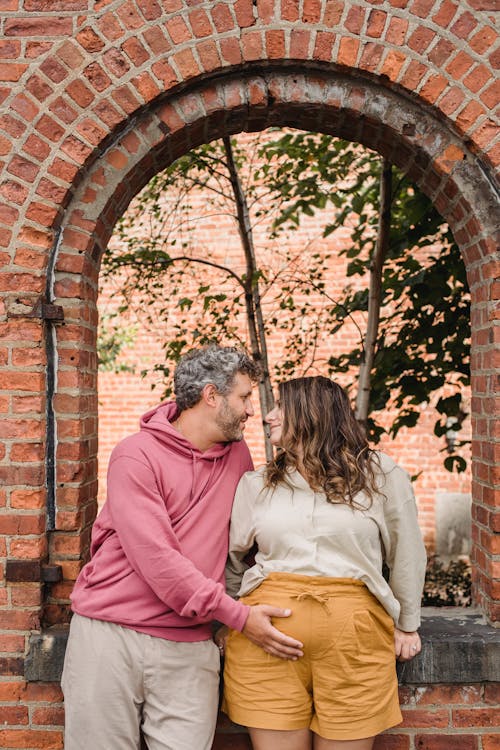 Stylish adult man in casual clothes touching tummy of pregnant wife while standing together near brick house and looking at each other