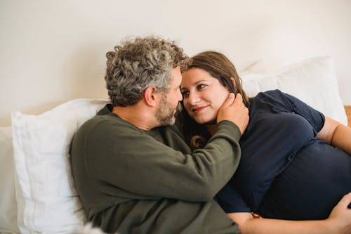 Enamored adult man in casual clothes lying on bed and touching face of happy pregnant wife during weekend at home