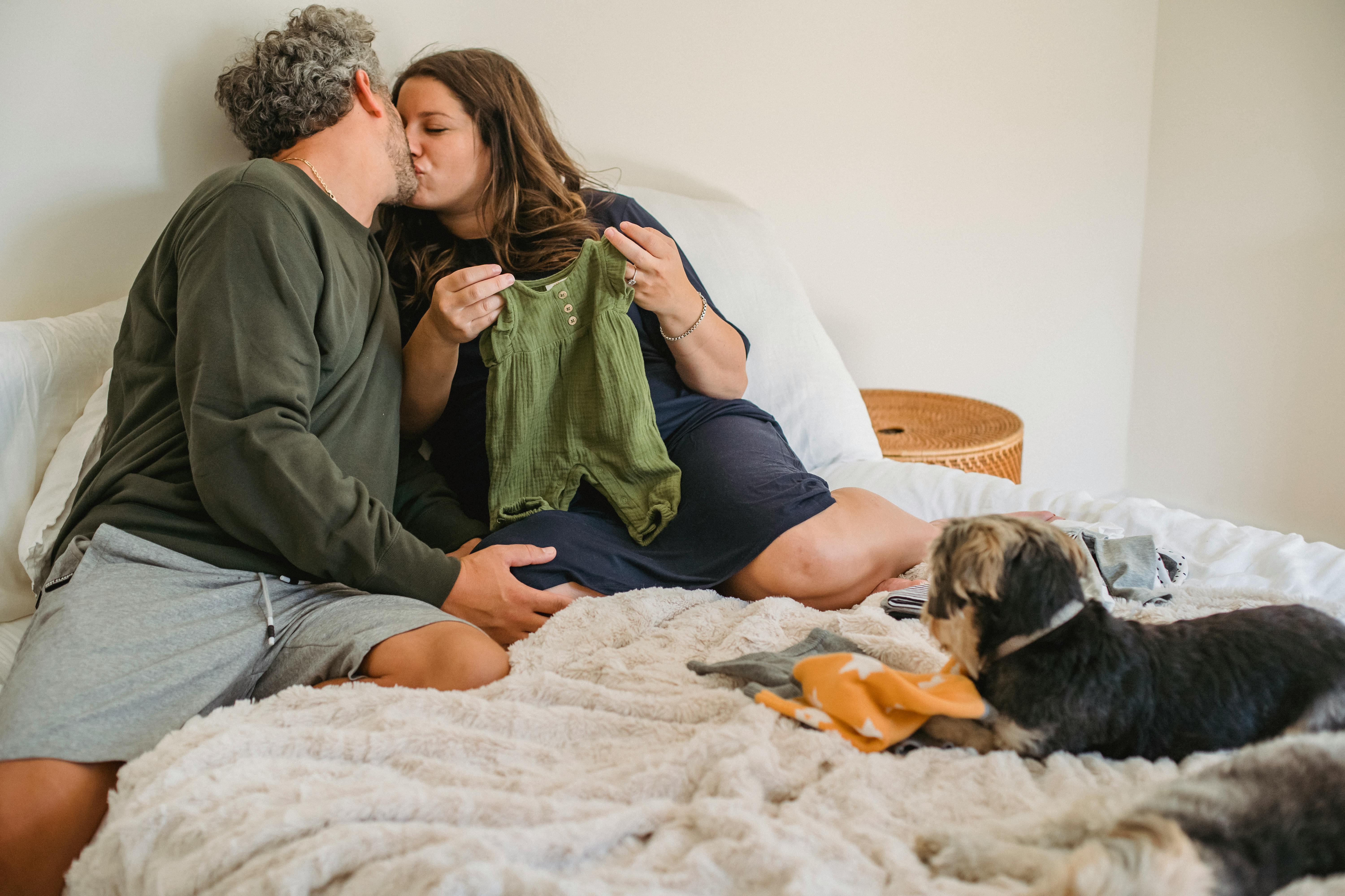 delighted pregnant couple kissing while relaxing on bed with puppies