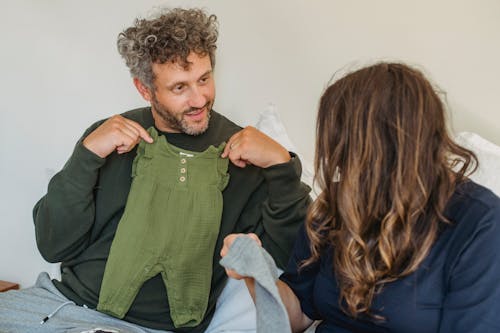 Free Happy adult father showing cute baby clothes to pregnant wife while resting together on bed at home Stock Photo