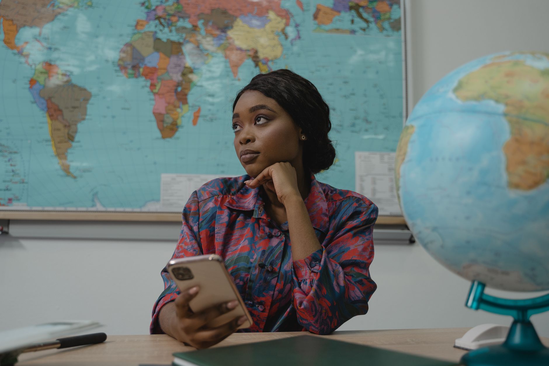 Best Phone Setup for Teaching Classes on Your Phone A teacher in a classroom, pondering with a smartphone. Educational setting with a globe and map.