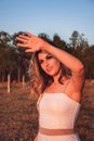 Elegant smiling female in white outfit standing on lawn near lush forest and raising arm to hide eyes from blinding sunlight
