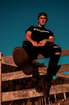 From below of young trendy male in stylish outfit and cap sitting on wooden fence in countryside against cloudless blue sky at sundown