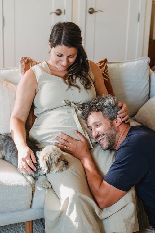 Smiling man listening to belly of pregnant wife resting on sofa with pet