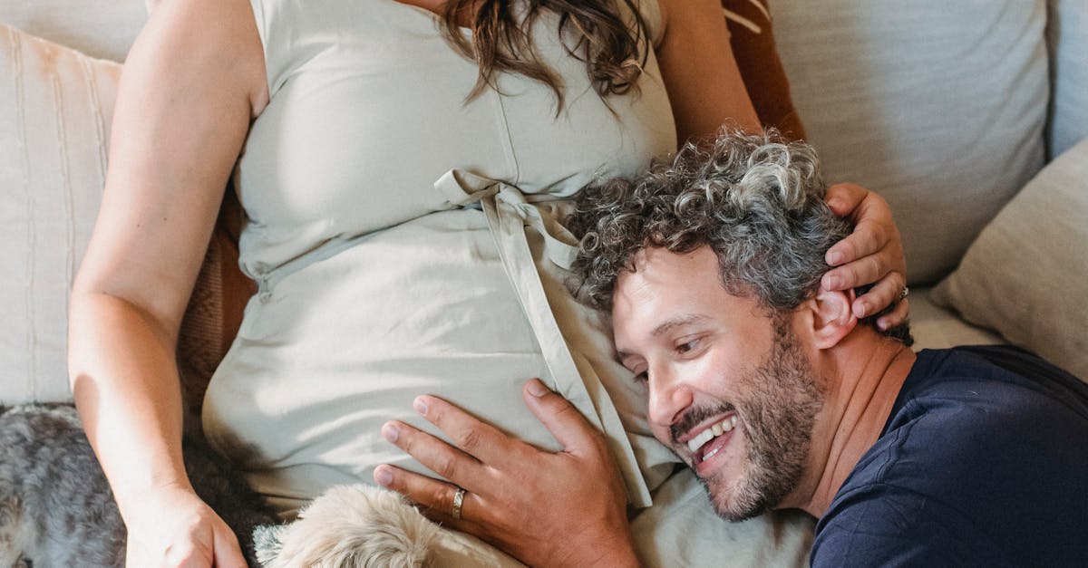 Smiling man listening to belly of pregnant wife resting on sofa with pet