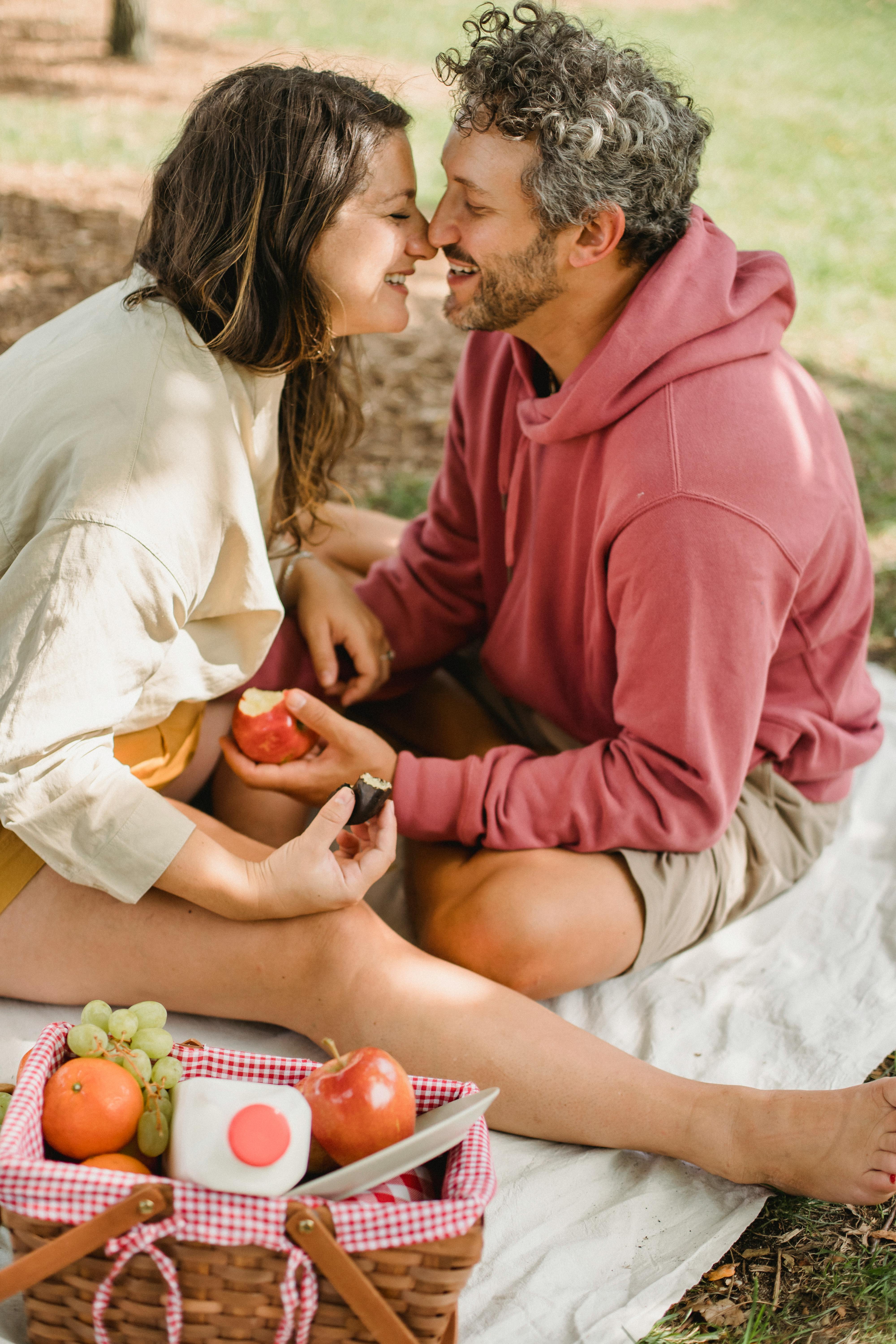 delighted romantic couple smiling and eating fruits during picnic in park