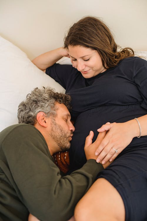 Free High angle of bearded male touching and kissing tummy of pregnant wife with closed eyes while lying together on comfortable bed in morning Stock Photo