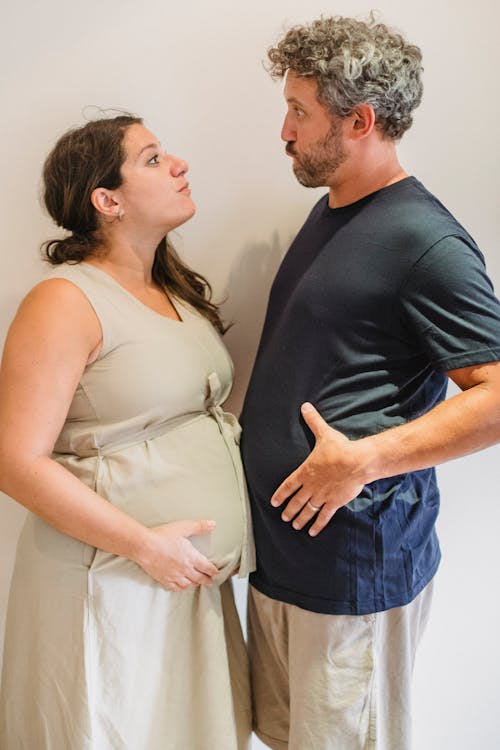 Side view of joyful adult man and pregnant woman in casual clothes comparing bellies and making grimaces while having fun together against white background