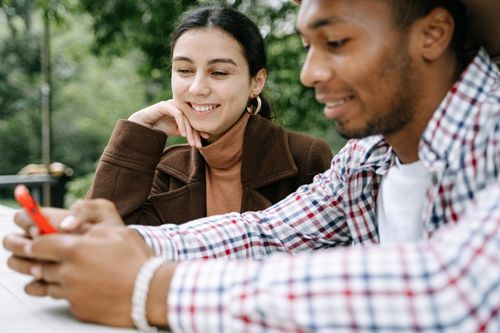 Free Two friends smiling and enjoying time together outdoors while using a smartphone. Stock Photo