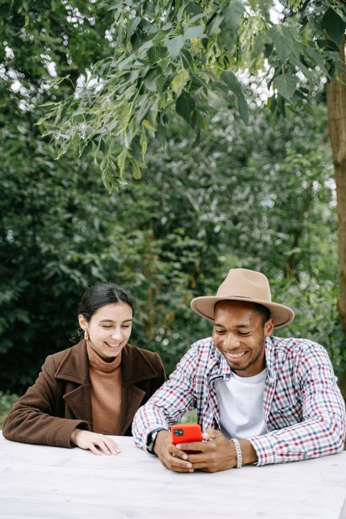 Free Two friends sitting at a table outdoors, smiling and looking at a phone, enjoying leisure time together. Stock Photo