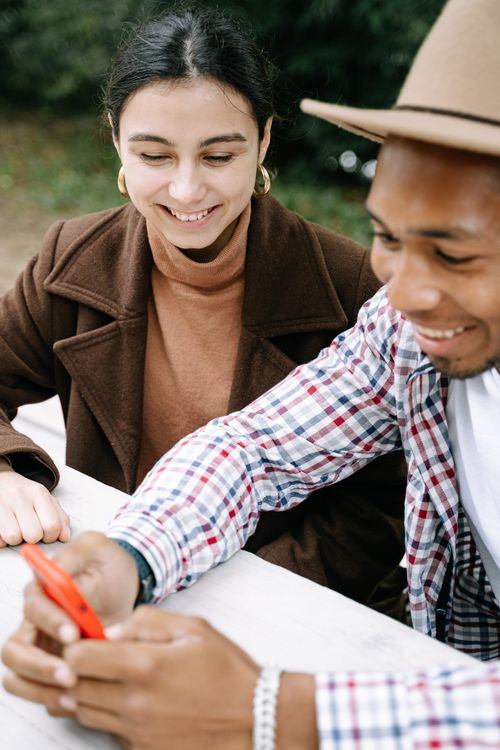Free Two friends sitting outdoors, smiling and looking at a smartphone together. Stock Photo