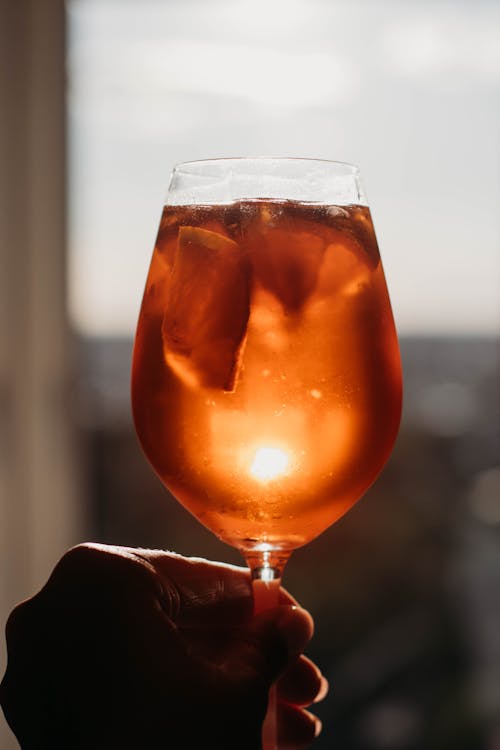 Crop anonymous person raised hand with orange cocktail with ice cubes in glass against window in daytime