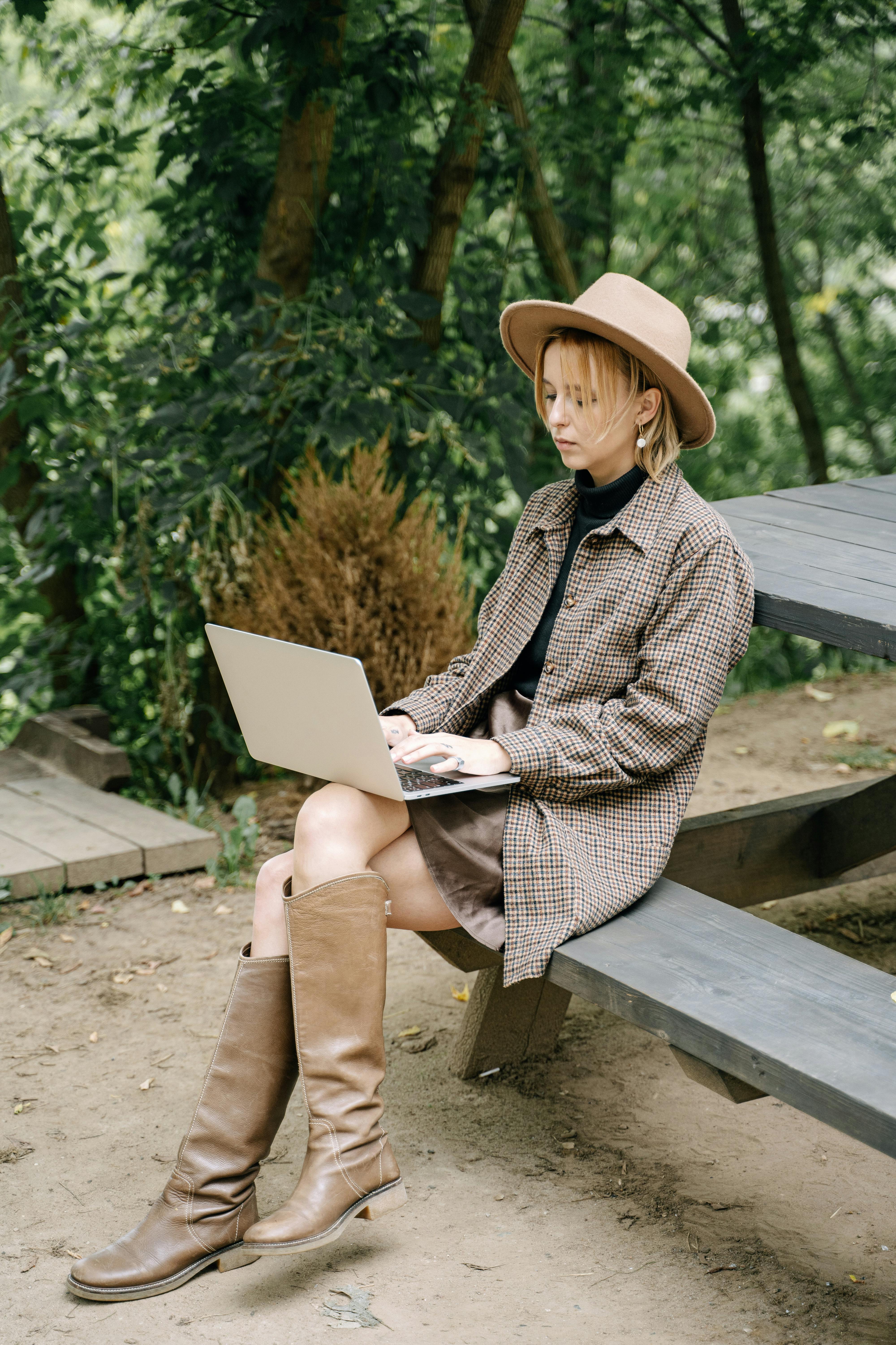 Woman working on a laptop outdoors in a park wearing a stylish hat and boots.