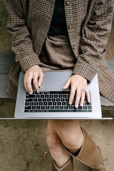 A woman in a brown coat typing on a laptop while sitting on a wooden bench outdoors.