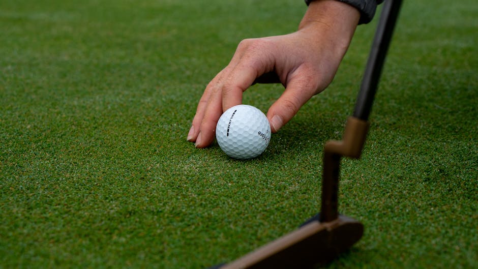Hand placing a golf ball on green grass with putter nearby.