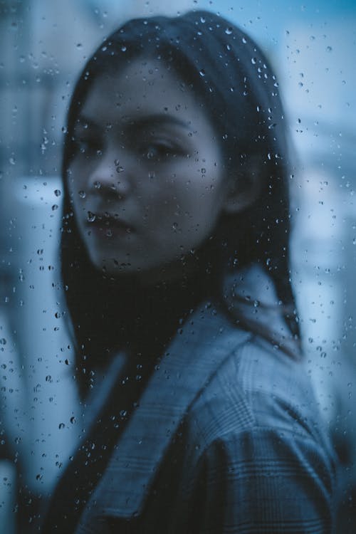 Through window side view of melancholic girl with dark hair thinking on problem in rainy day