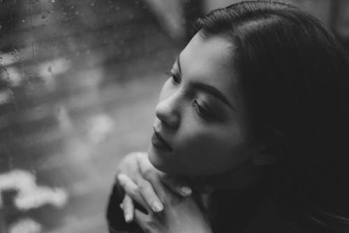 Black and white high angle of serious teenage girl sitting in solitude near window covered with raindrops