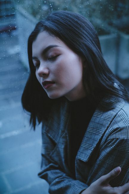 From above of tranquil girl with closed eyes contemplating while resting near window with waterdrops