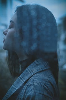 Through glass side view of upset teen girl with closed eyes standing behind wet window in rainy day
